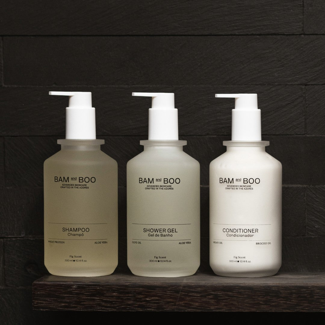 Three bottles of BAM and BOO shampoo, shower gel, and conditioner on a wooden shelf with a dark background. Natural Premium Amenities Portugal